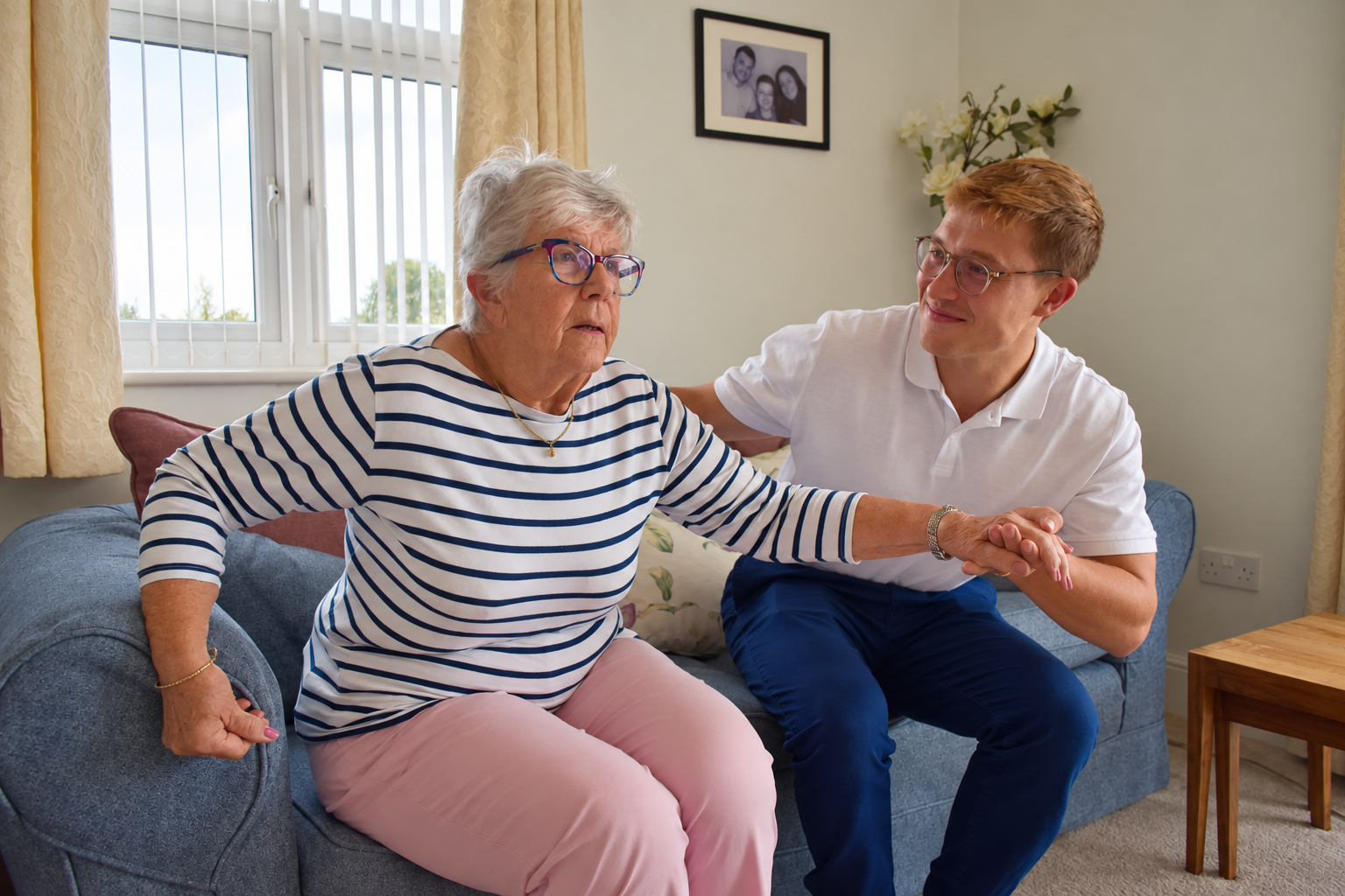 A physiotherapist supports an older woman as she gets up from a sofa at home.