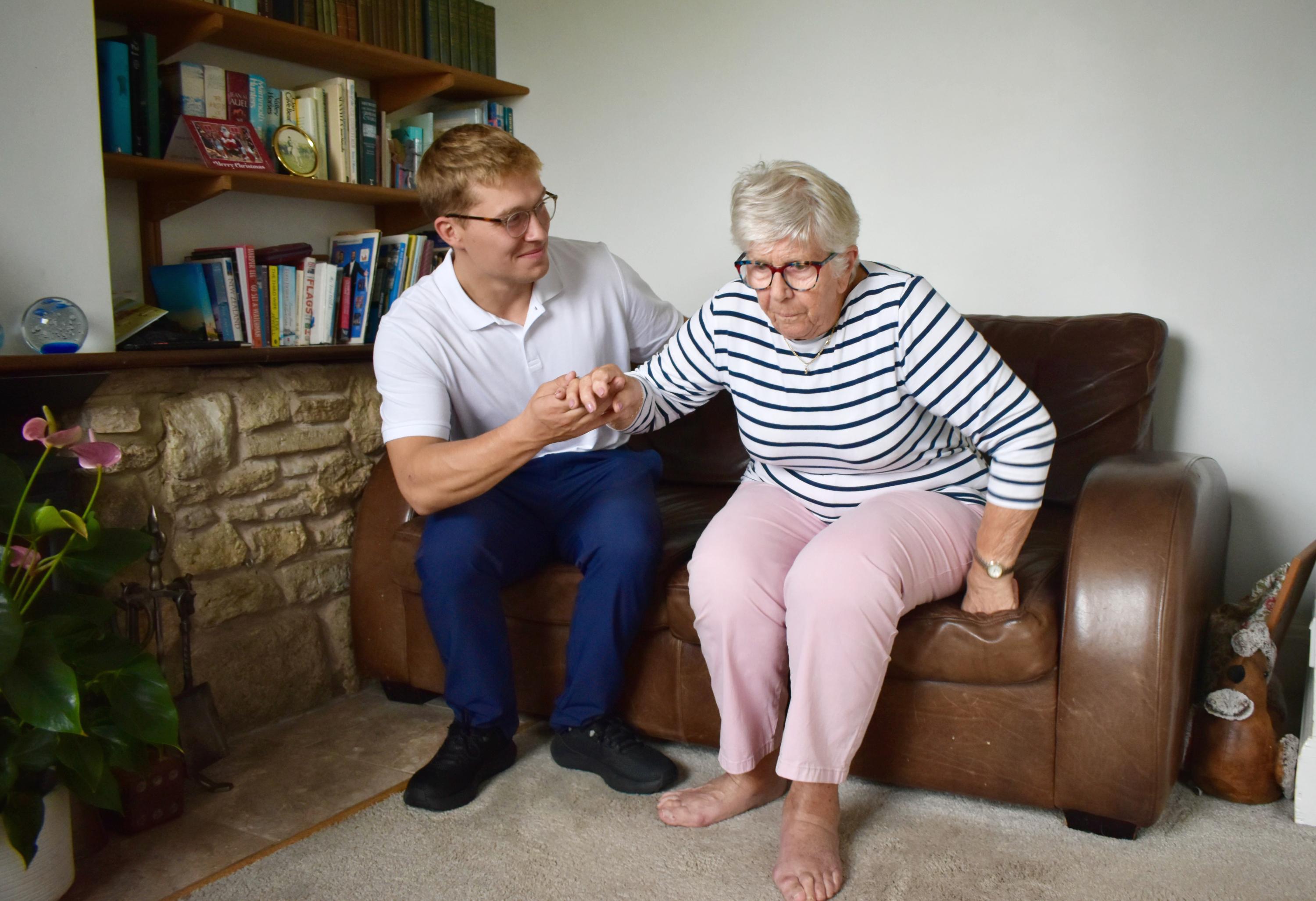 Jacob supports an older woman as she prepares to stand from her own sofa.